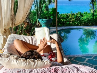 persona acostada en una cama frente a una piscina con vista al mar mientras lee un libro