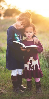 Dos niñas están de pie, la más alta de ellas tiene el brazo recargado en la niña menor que sostiene un libro en sus manos
