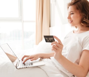 mujer sentada en la cama sostiene una tarjeta de crédito en una cama mientra utiliza su laptop