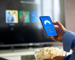 Persona sostiene un celular en la mano con la imagen de una VPN y al fondo se observa una televisión