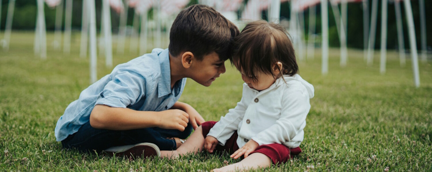 Dos niños pequeños, probablemente hermanos, sentados en la hierba y unidos frente a frente