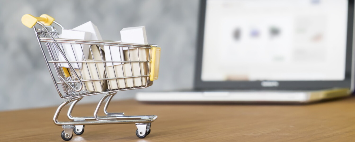 Carrito de la compra en miniatura con cajas sobre una mesa con un pc en el fondo