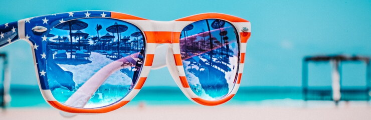 A pair of sunglasses with American Flag frames reflect beach chairs and umbrellas in the lenses