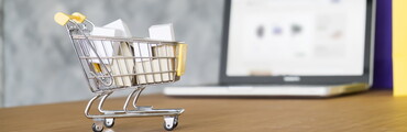 Carrito de la compra en miniatura con cajas sobre una mesa con un pc en el fondo