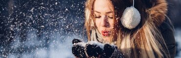 girl with earmuffs, heavy coat and gloves blowing snow out of her hands