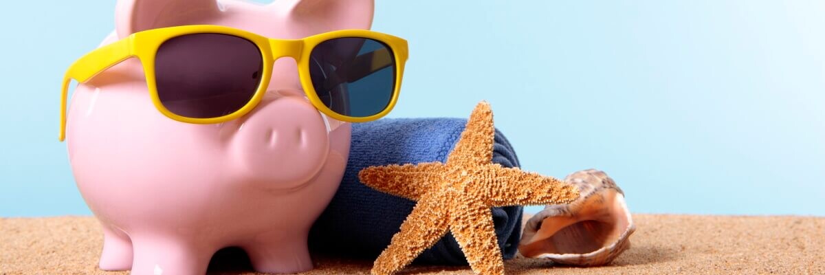 Alcancía con forma de cerdito rosa, con gafas de sol amarillas sobre la arena de la playa, con toalla, estrella y concha de mar
