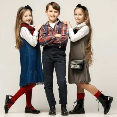 Un garçon en salopette et chemise à carreaux à côté de deux filles aux cheveux longs et chaussettes rouges : achetez des vêtements pour vos enfants chez Kids Around avec le code de réduction bravopromo.fr