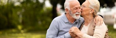 Señor y señora mayores sentados en un parque, abrazados y ella dándole un beso a él en la mejilla.