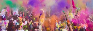 Crowd celebrating Holi festival with colorful powders in the air, hands raised and joyful atmosphere.